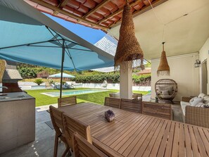 An outdoor patio with a pool, wooden furniture, and a pergola with a blue canopy. - Villa Claudia by Hello Homes Sitges (Sitges)