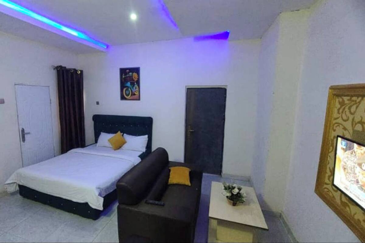 Un dormitorio con cama, sofá, una mesita con un jarrón, una puerta y un televisor montado en la pared.