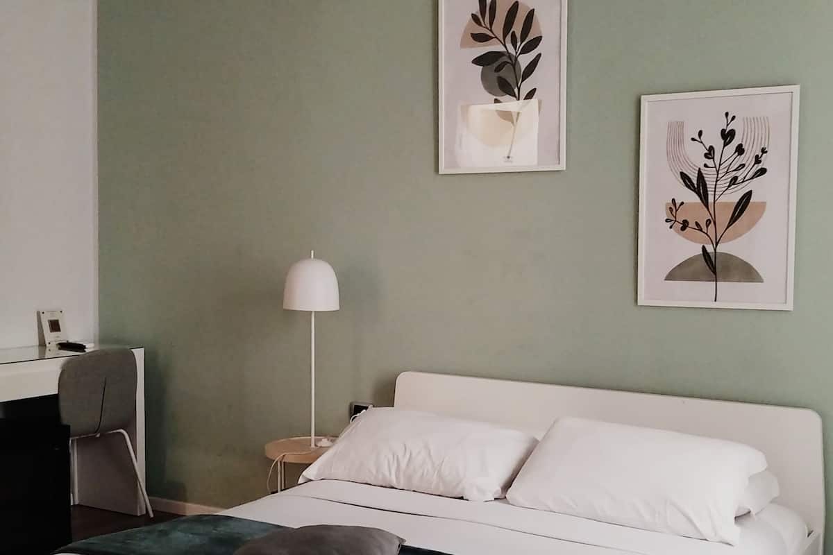 A neatly made bed with white bedding and a teal throw, a white lamp beside it, and two framed botanical prints on the wall.