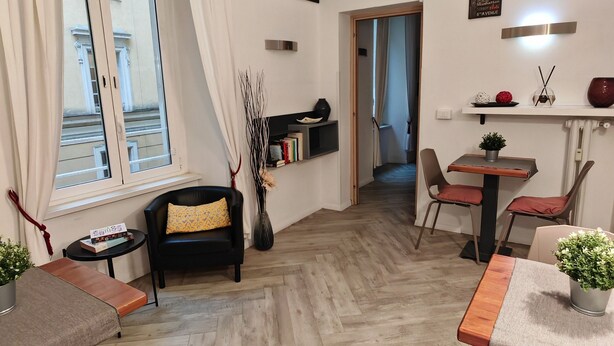Un salon moderne avec un parquet en chevrons, un canapé noir agrémenté d’un coussin jaune, une petite table ronde avec un vase, et un coin repas avec un banc et des chaises en bois.