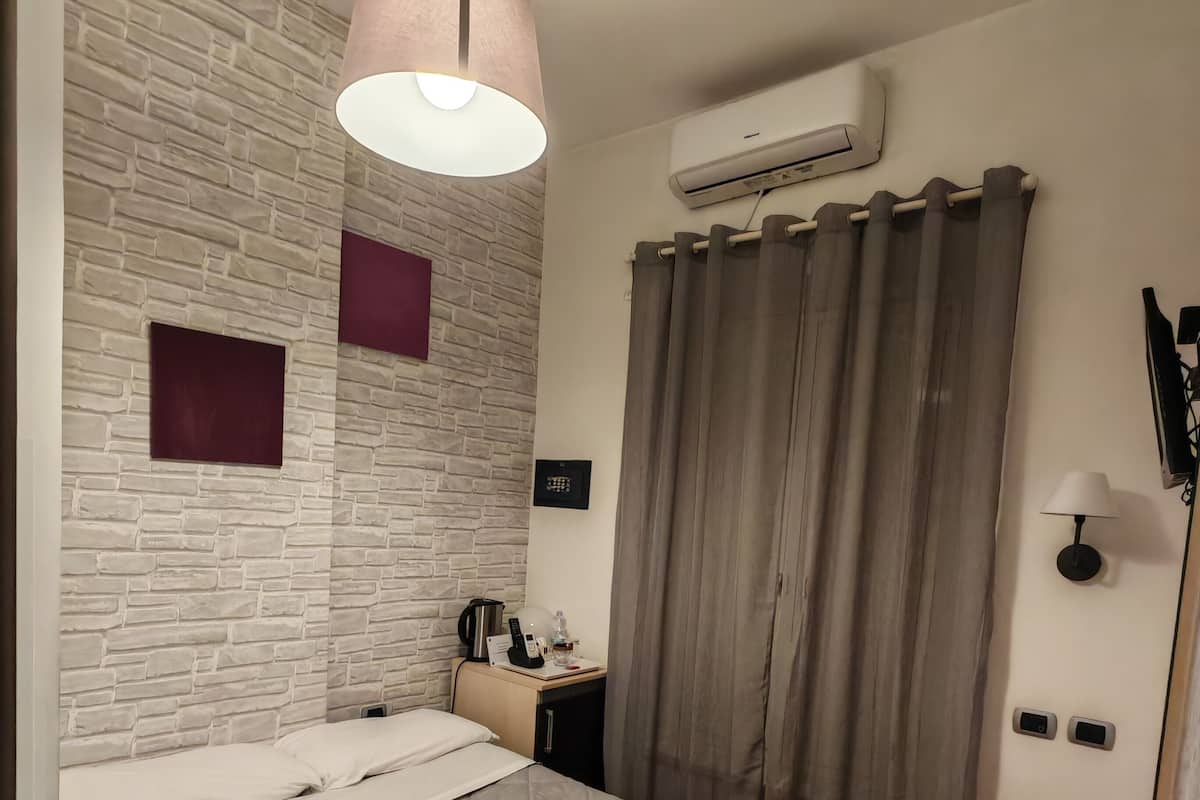 A modern hotel room with a brick-patterned wall, a bed with an orange blanket, a bedside table with a lamp, and an air conditioning unit.