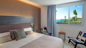A modern hotel room with a large bed, a seating area with chairs, a small table with fruit, and a view of the sea through large windows.