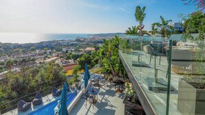 Una terrazza sul tetto con piscina, aree salotto e ringhiera in vetro che si affaccia su una vista panoramica della città e del mare.
