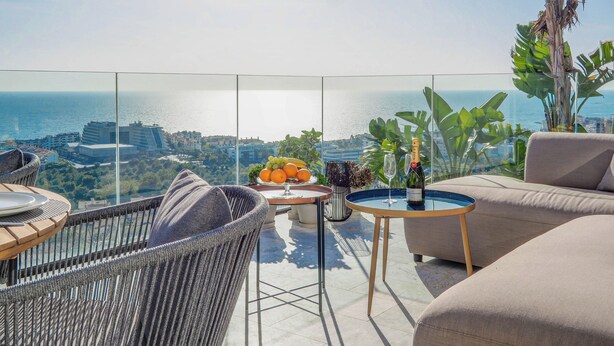 Un balcone con vista sul mare, arredi in vimini, un tavolo con frutta e una bottiglia di champagne, e una pianta in vaso.
