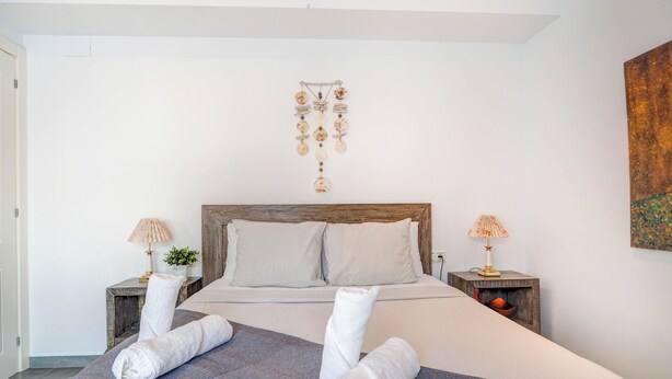 A neatly made bed with a grey blanket and two rolled towels, a wooden headboard, two bedside tables with lamps, and a wall art piece above the bed. - Central Terrace by Hello Homes Sitges (Sitges)