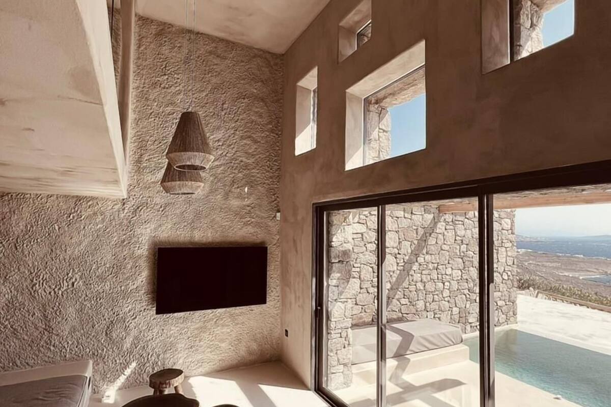 A modern interior with a stone wall, a flat-screen TV, a large window with a view of the sea, and a minimalist seating area.