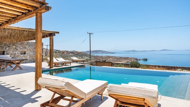 Uma área à beira da piscina com espreguiçadeiras, um muro de pedra e vista para o mar.