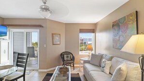 A living room with a glass table, a sofa, a chair, a ceiling fan, a window with a view, and a painting on the wall. - Grand Caribbean 325 by Liquid Life (Orange Beach)