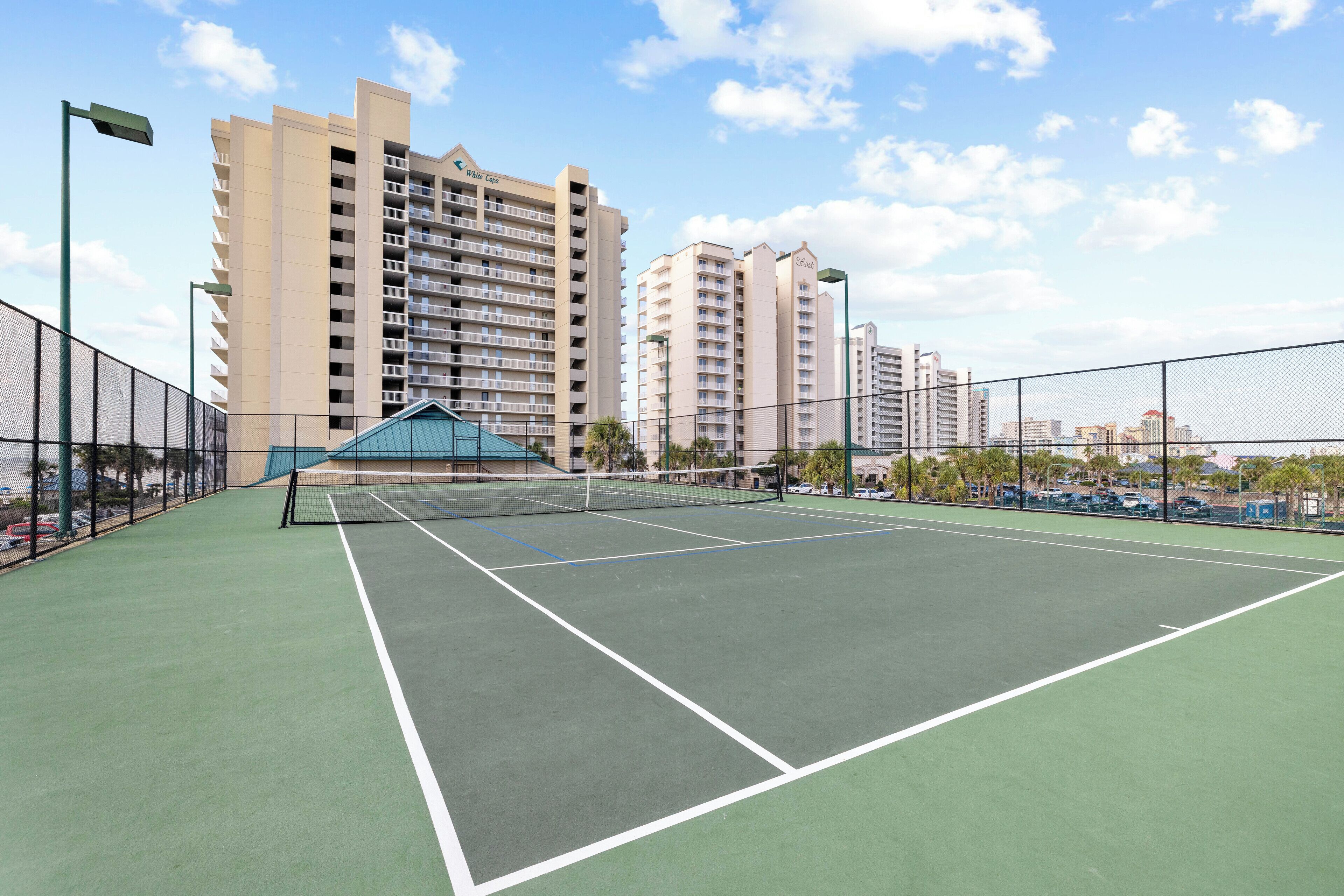 An outdoor tennis court with a net, surrounded by a fence, and adjacent to tall residential buildings.
