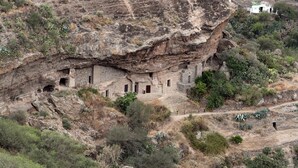 Un acantilado con antiguas viviendas excavadas en la roca, rodeado de vegetación escasa y un sendero de tierra.