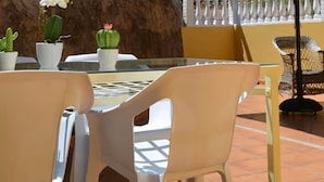 Un patio con sillas de plástico y una mesa, rodeado de plantas en macetas y una valla blanca.