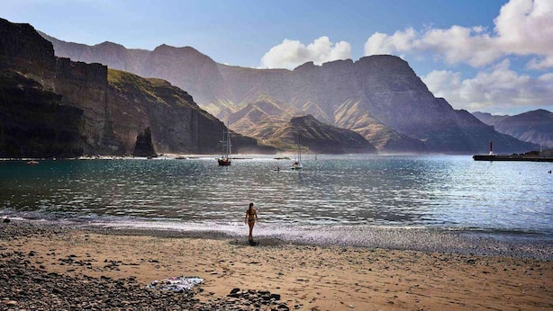 Una playa con terreno rocoso, una persona de pie y barcos en el agua.