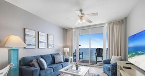 A living room with a sofa, coffee table, and TV, a ceiling fan, and a balcony with a view of the ocean. - Lighthouse 1206 by Liquid Life (Gulf Shores)