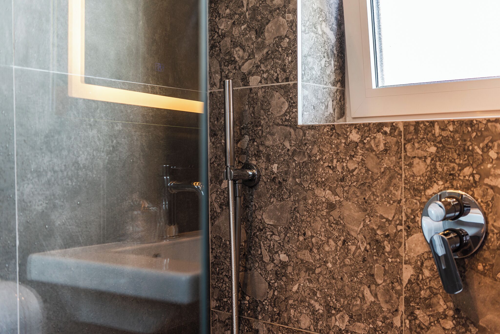 A bathroom with a glass shower enclosure, a sink, and a marble-tiled wall.