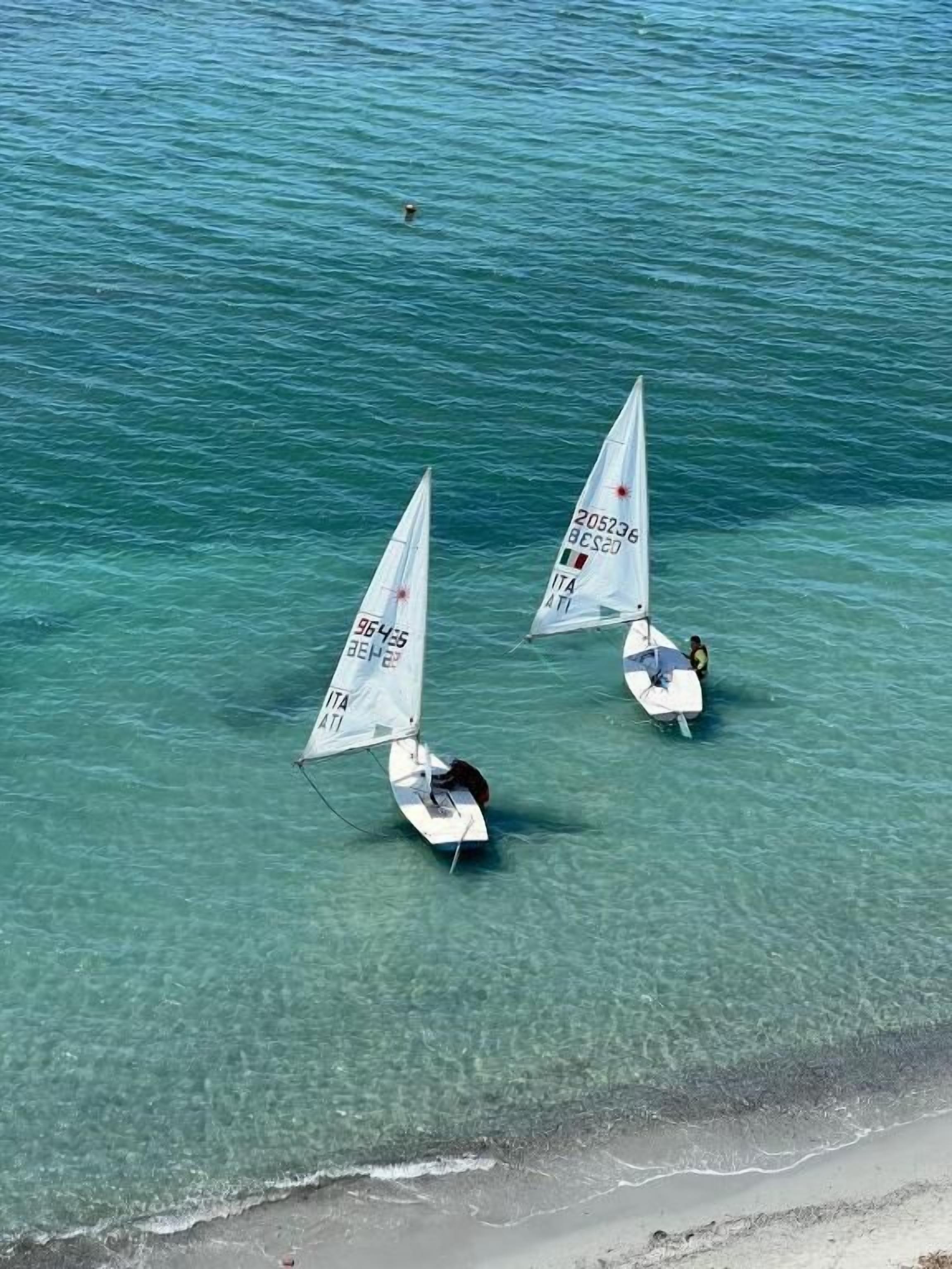 Twee zeilboten met ‘ITA’ op de zeilen op een rustige zee nabij een strand.