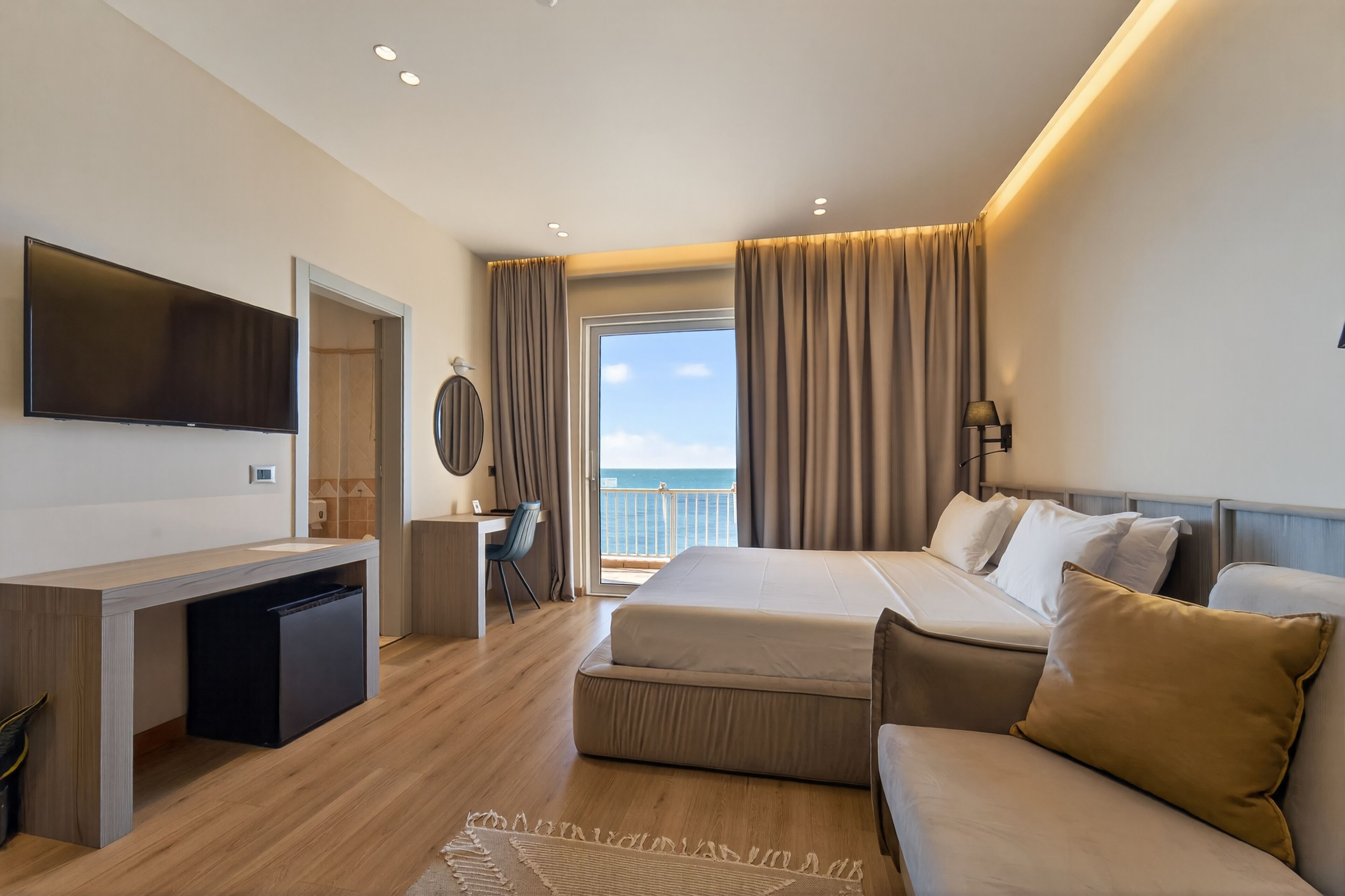 A modern hotel room with a large bed, a desk, a flat-screen TV, and a view of the sea.