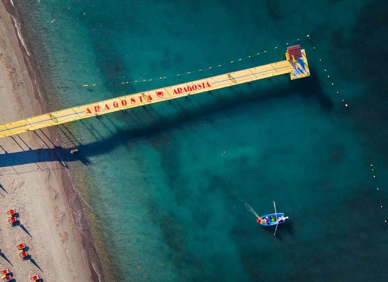 Vista aérea de una playa con un muelle identificado como 'ARAGOSTA' que se adentra en el agua, un bote pequeño y varias personas en la orilla.