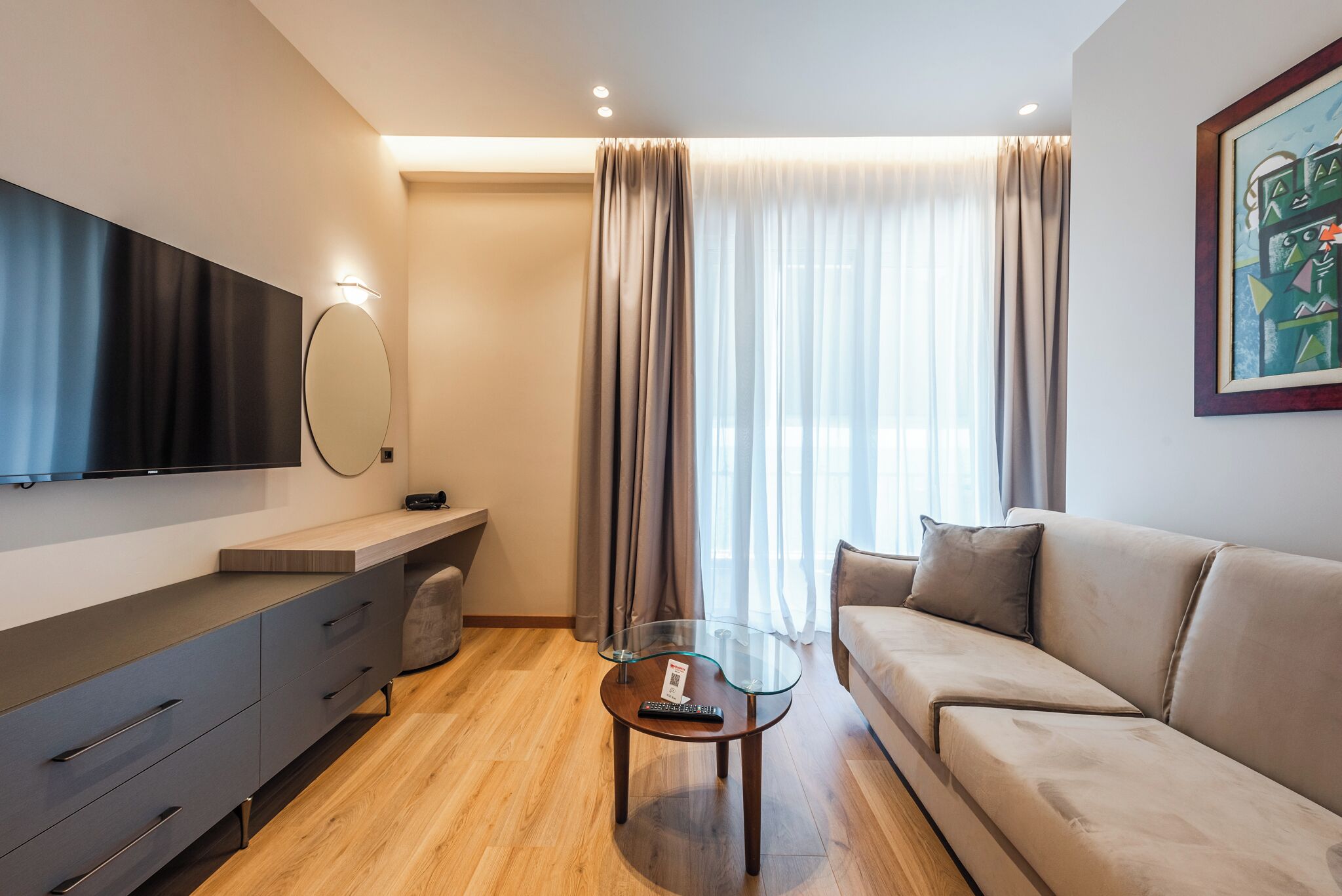 Ein modernes Wohnzimmer mit einer Couch, einem Glastisch, einem an der Wand montierten Fernseher und einem großen Fenster mit Vorhängen.