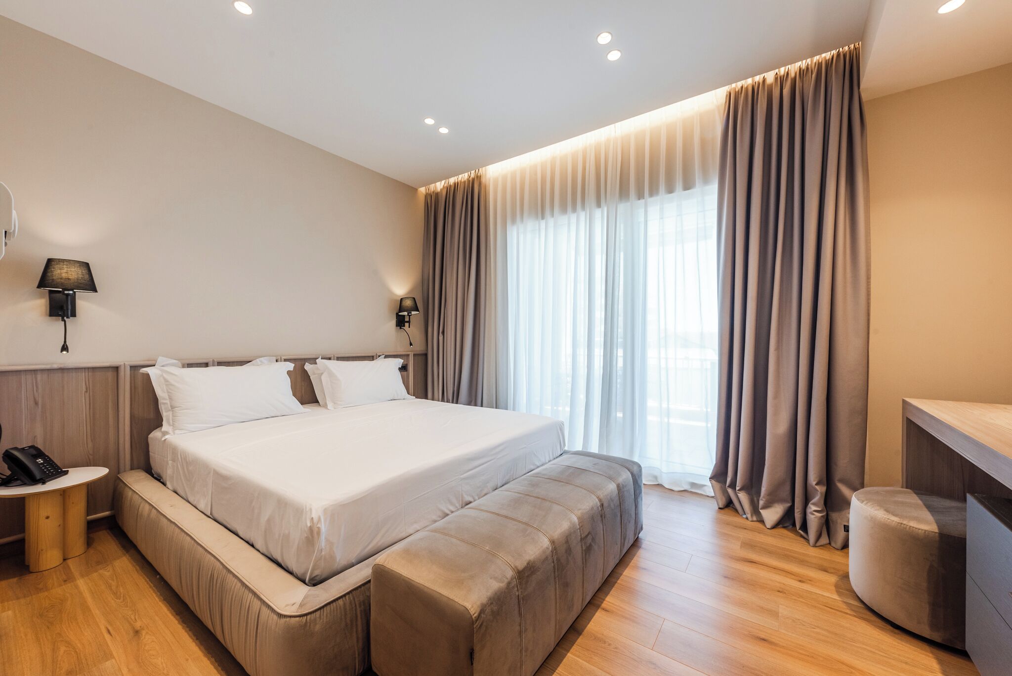 A modern hotel room with a large bed, bedside lamps, a desk with a telephone, and a large window with curtains.