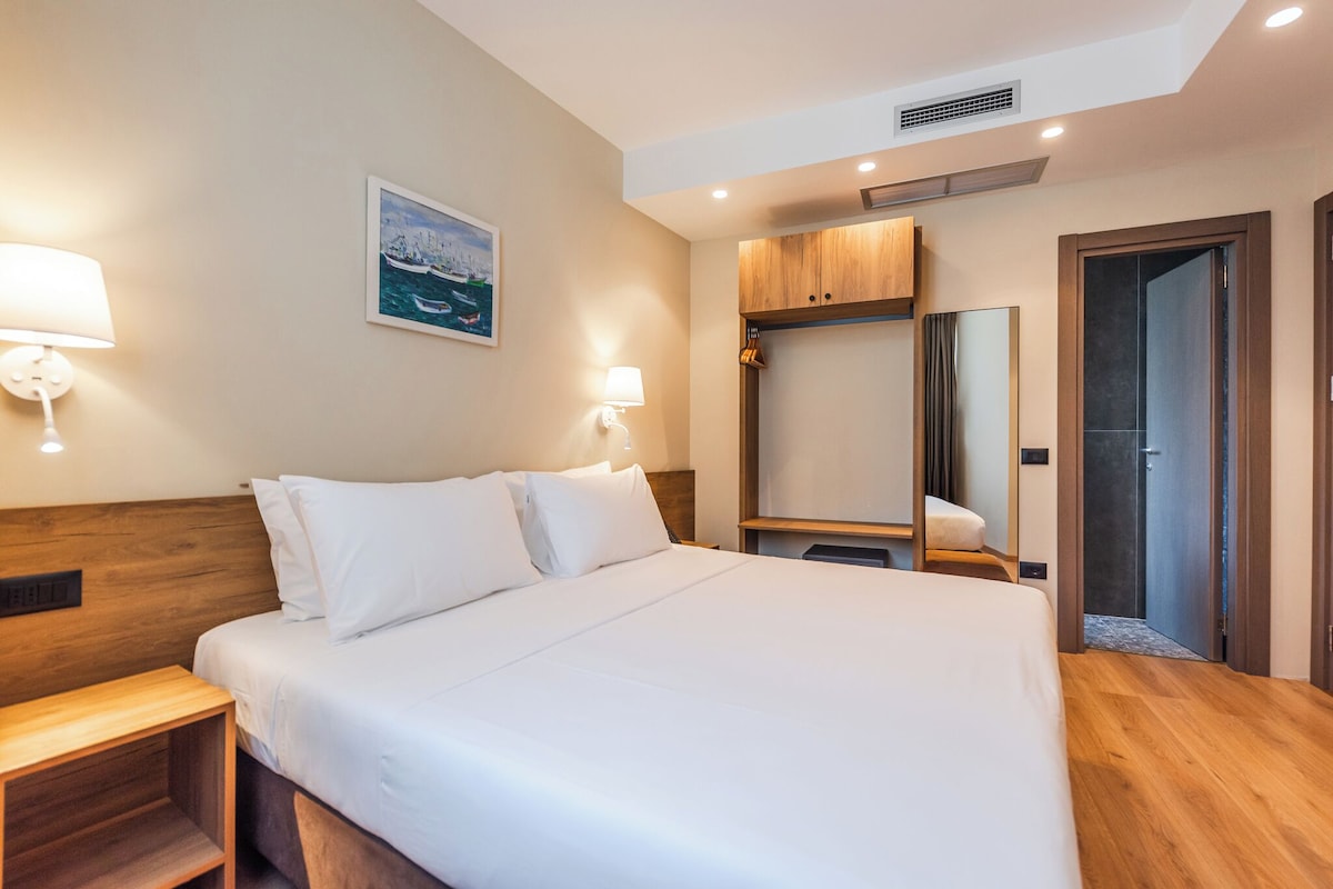 Habitación de hotel con una cama grande, cabecera de madera, dos lámparas de noche, una mesita de noche, un espejo y una puerta que da a un baño.