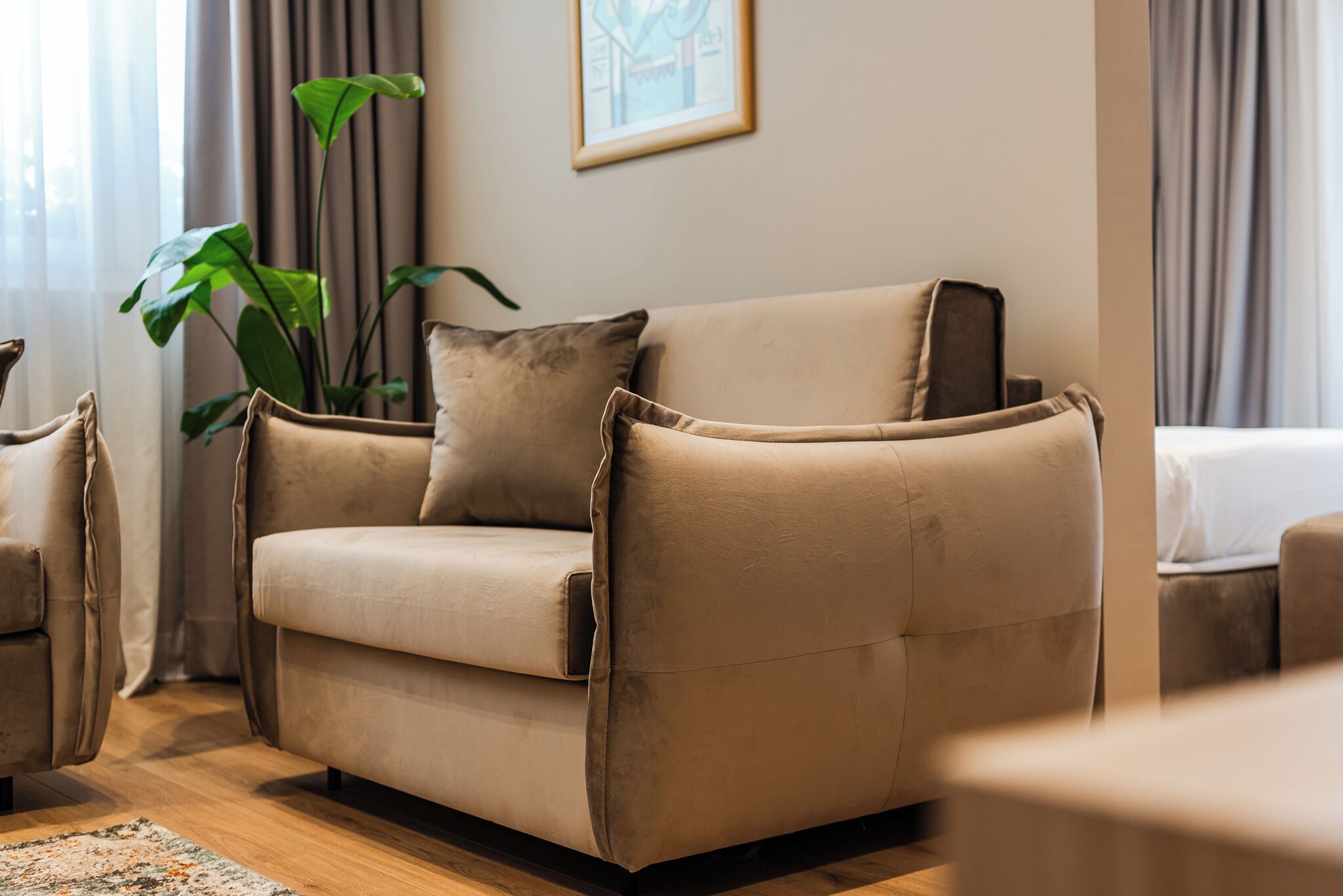 A beige sofa with a matching armchair and a small table in a well-lit room with a large plant and a framed picture on the wall.