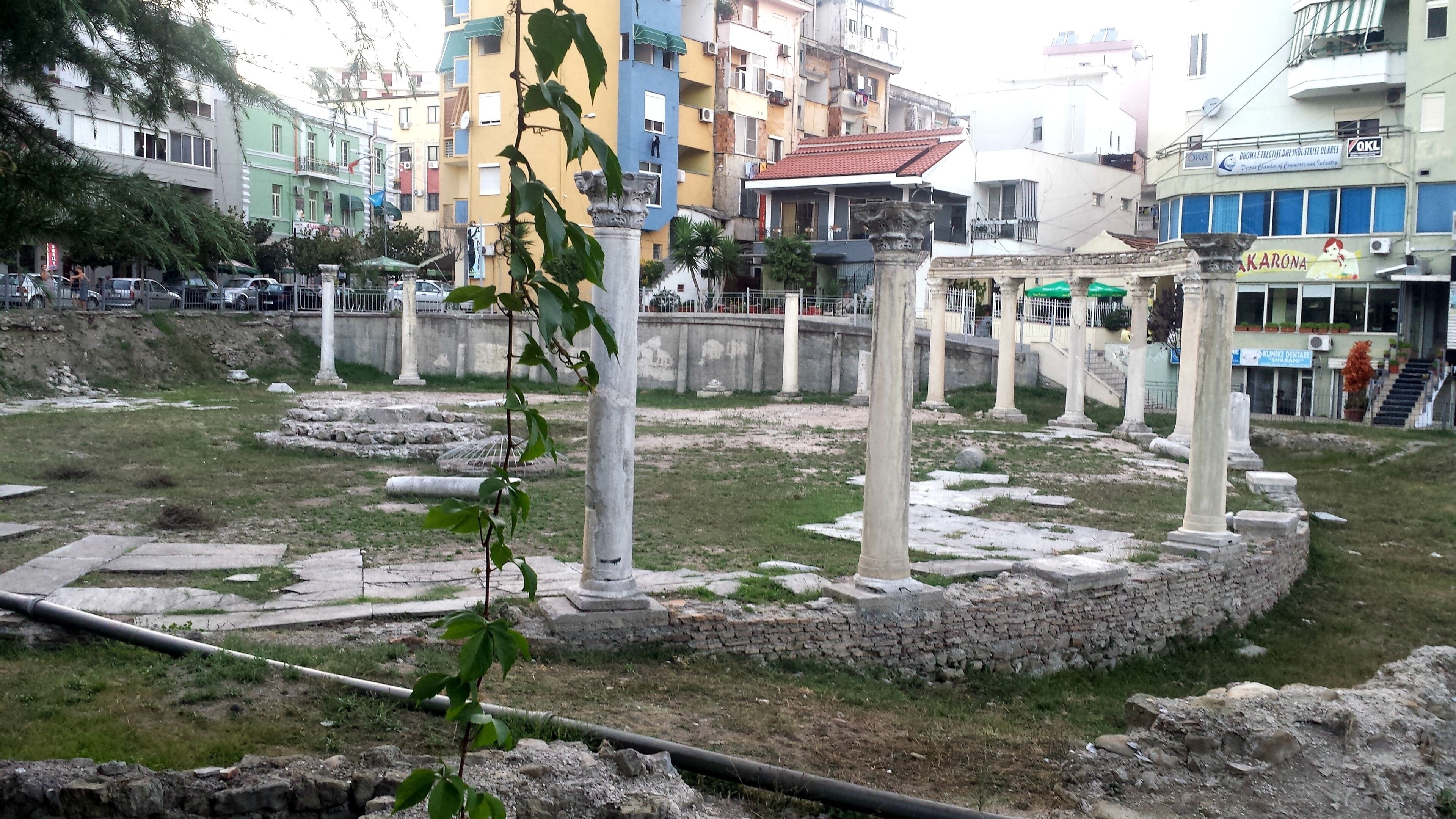 An archaeological site with ancient ruins, including columns and stone structures, surrounded by grassy areas and modern buildings in the background.