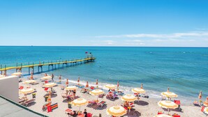 A beach with numerous sun umbrellas and lounge chairs, a pier extending into the sea, and a clear blue sky. - Aragosta Hotel (Durrës)
