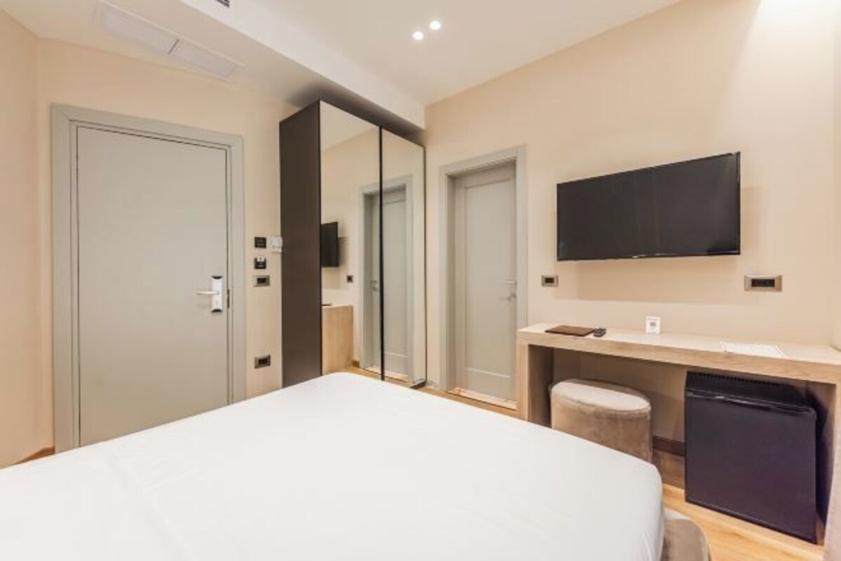 A modern hotel room with a bed, a TV mounted on the wall, a small desk, and a mini-fridge.