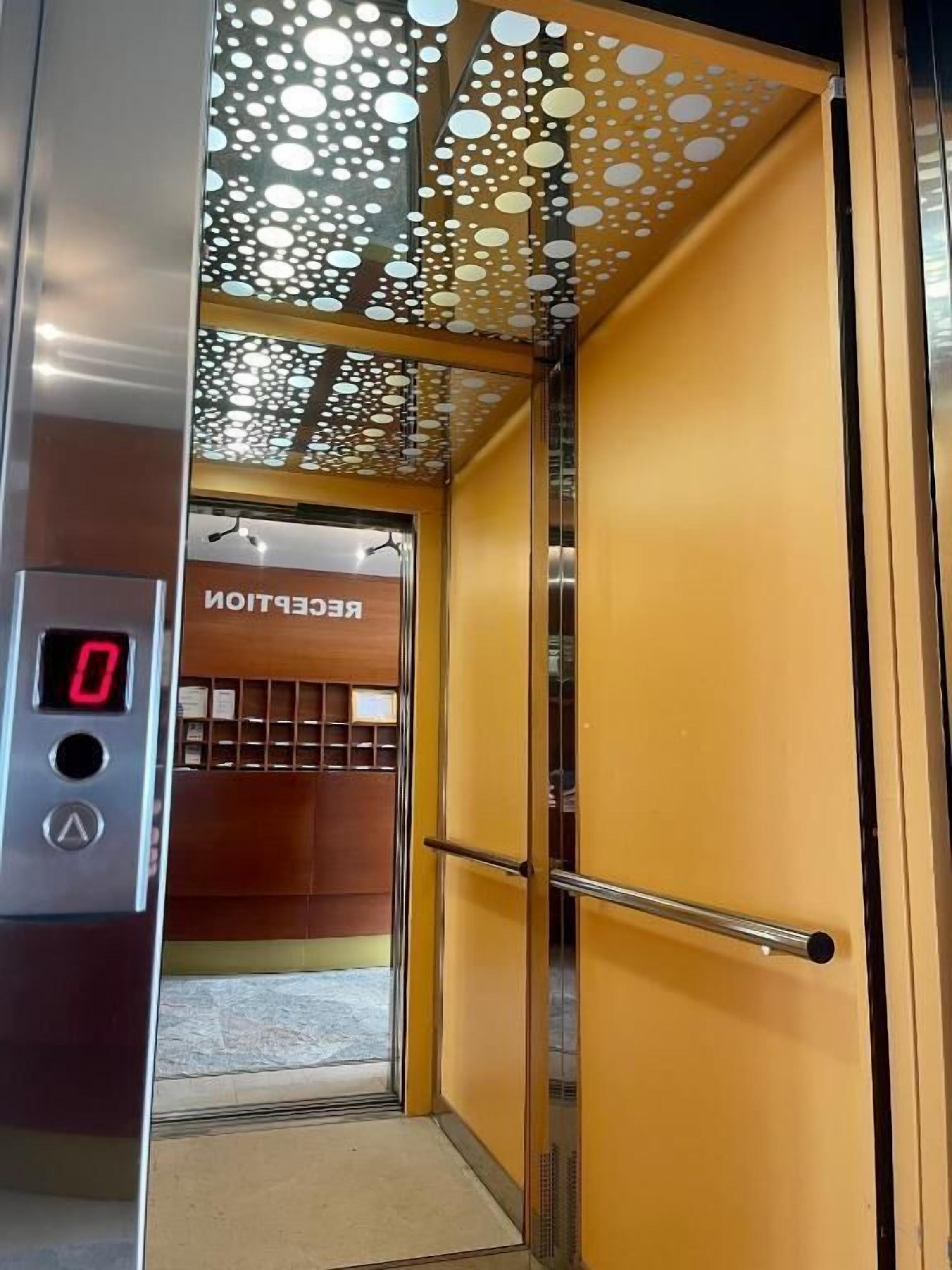 An elevator interior with a digital display, a metallic handrail, and a door leading to a reception area.