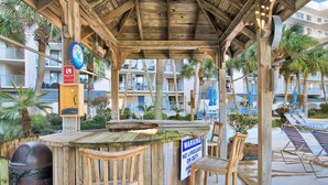 Un espace bar extérieur en bois, agrémenté d’un panneau indiquant la présence d’un sauveteur, de chaises au bord de la piscine et offrant une vue sur un immeuble à plusieurs étages doté de balcons.