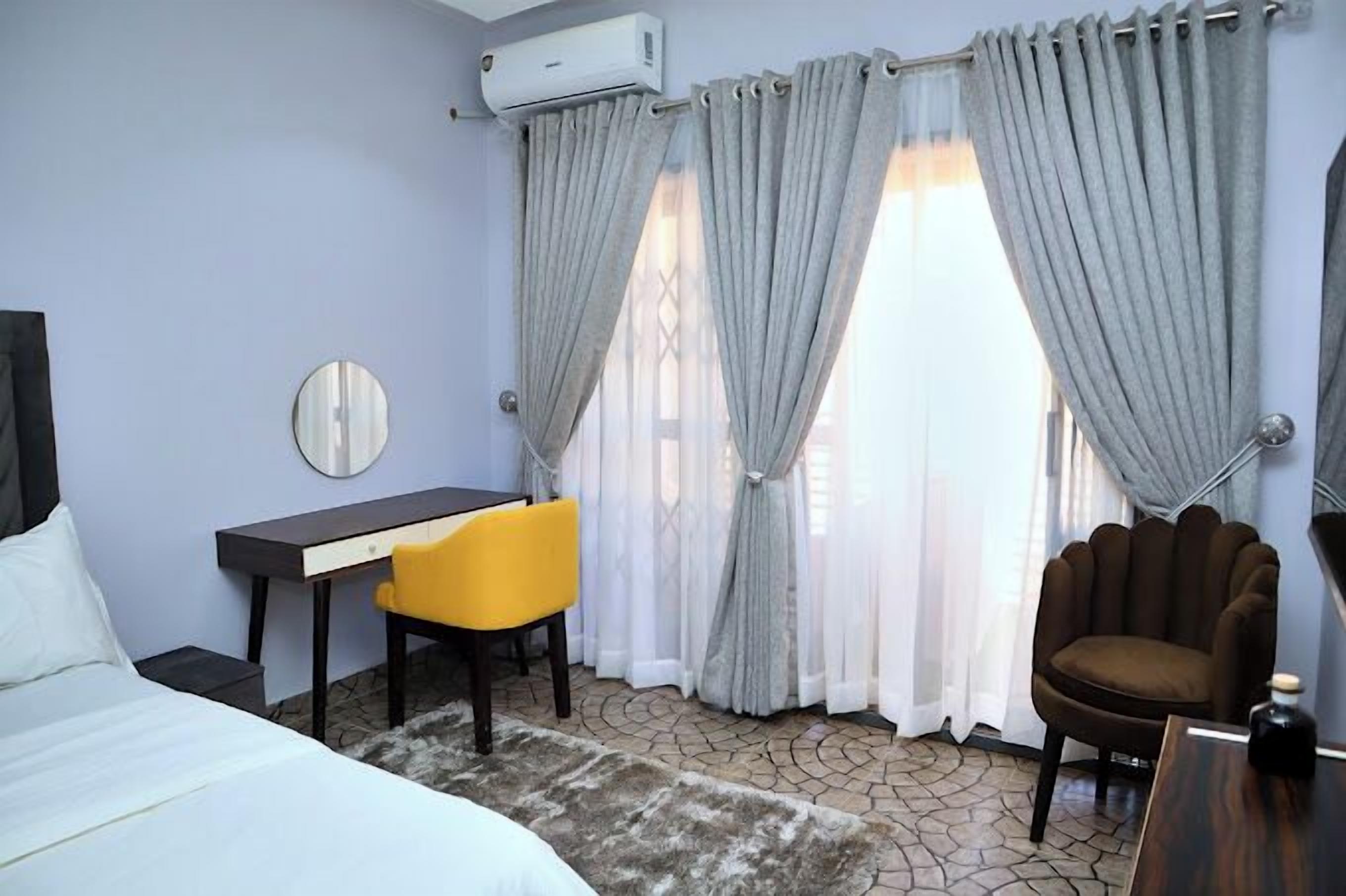A hotel room with a bed, a desk, a chair, a round mirror, and a window with curtains.