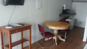 Een kleine, goed verlichte kamer met een houten tafel, een rode stoel, een aan de muur bevestigde televisie en een magnetron op een plank.