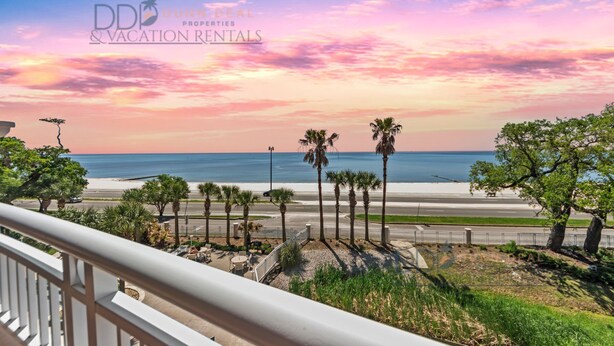 A balcony with a view of a beach, palm trees, and a sunset. - Legacy Tower II 305 | Stunning Coastal Views! (Gulfport)