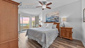 A bedroom with a bed, a ceiling fan, a nightstand, a lamp, a painting, and a view of the outdoors. - Legacy Tower II 305 | Stunning Coastal Views! (Gulfport)