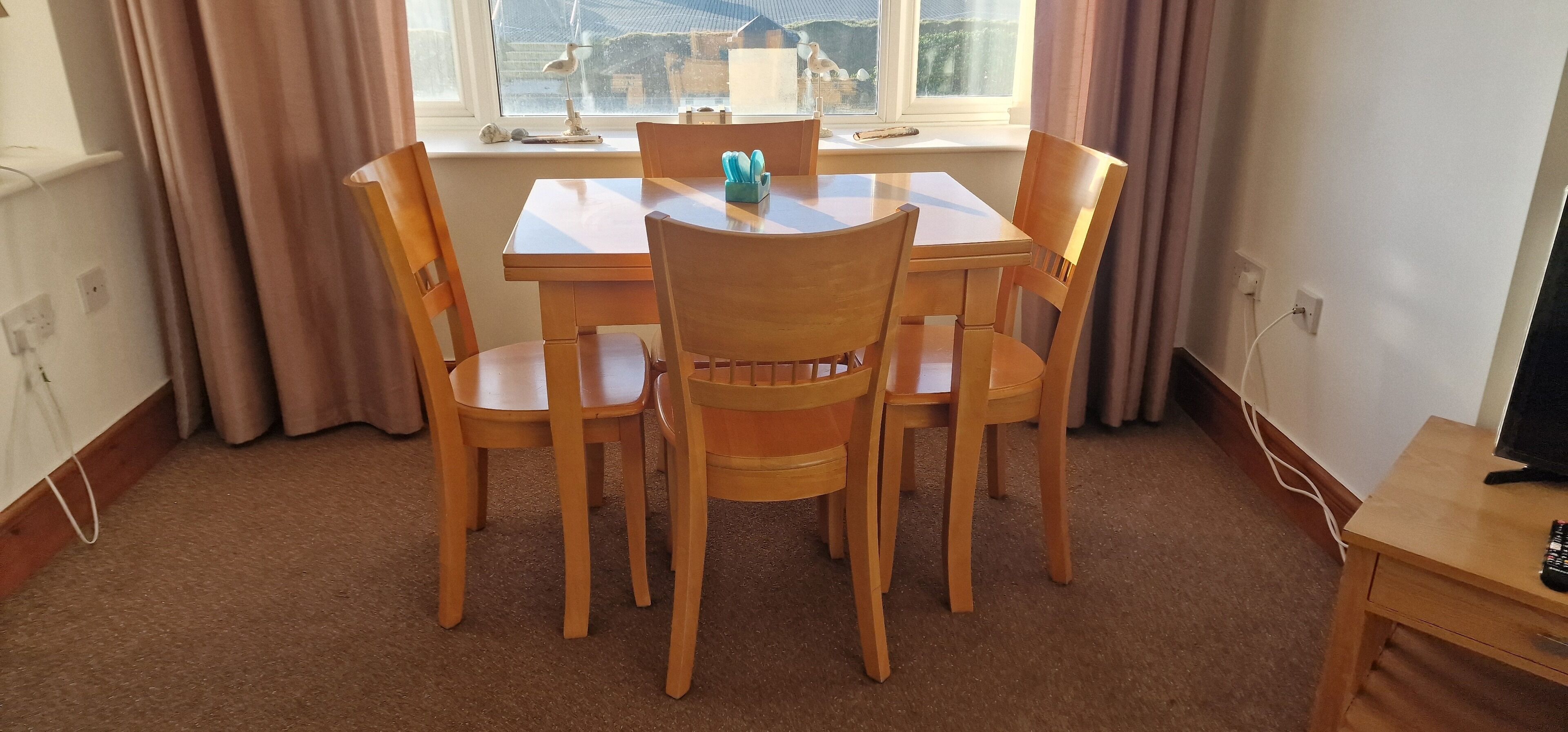 Un coin repas comprenant une table et quatre chaises, une fenêtre avec des rideaux et une petite table sur laquelle se trouve une télécommande.