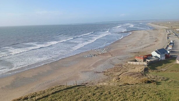 Vista costera con una playa arenosa, una colina cubierta de hierba y una hilera de edificios a lo largo de la orilla.