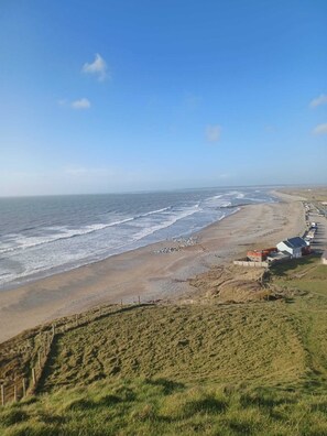A coastal view with a sandy beach, a grassy hill, and a row of buildings along the shore.