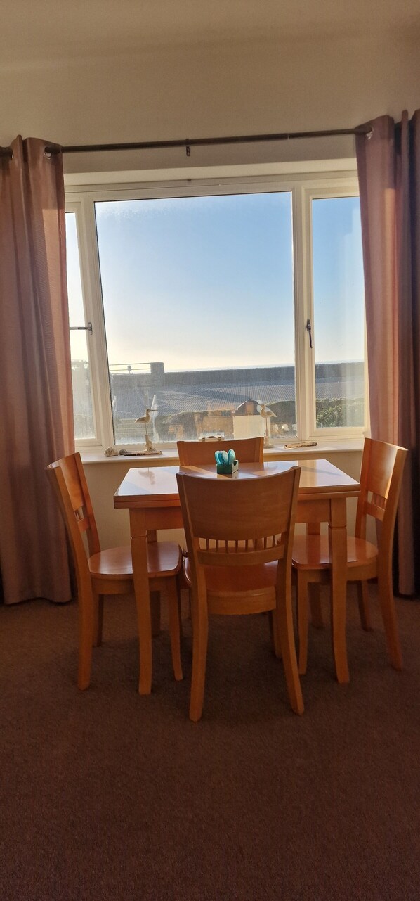 Una habitación con una mesa y sillas de madera, un ventanal con vista al mar y una cortina a la izquierda.
