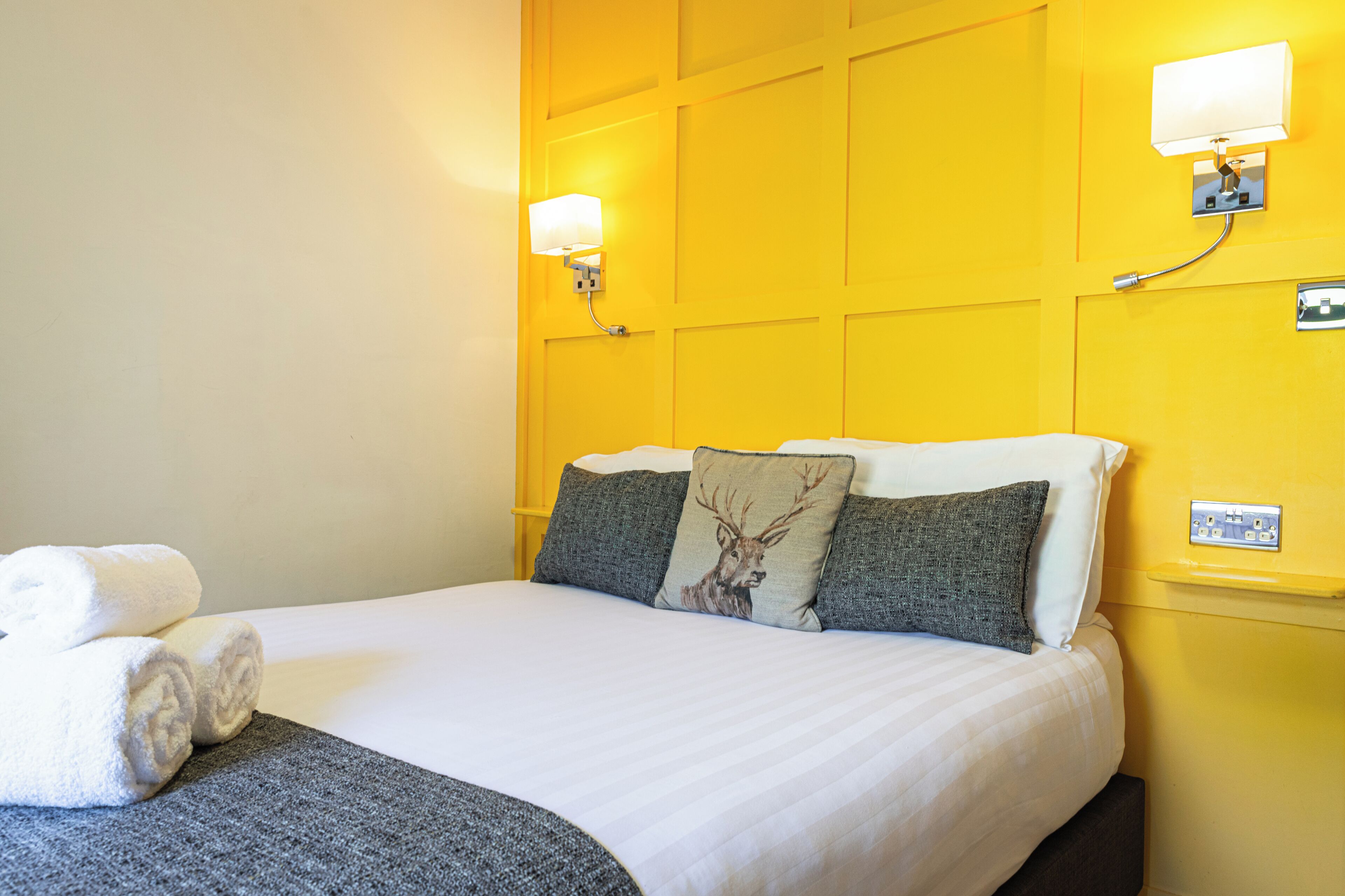 Una habitación de hotel con cama, almohadas, mesita de noche e iluminación en la pared.