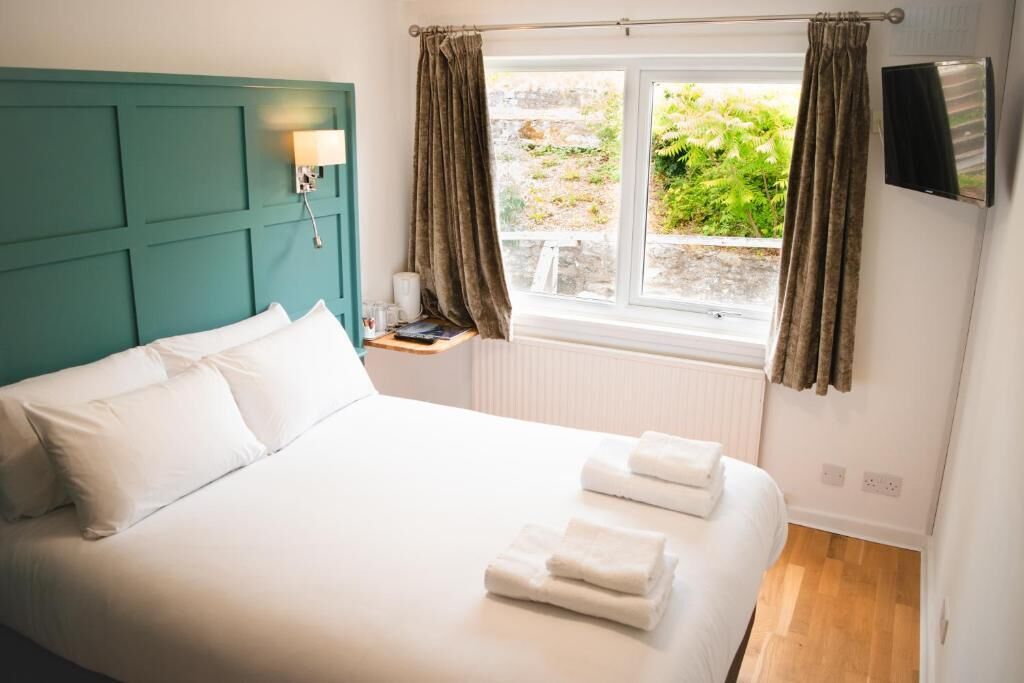 A neatly made bed with white linens and towels, a teal headboard, a bedside table with a lamp, and a window with curtains.