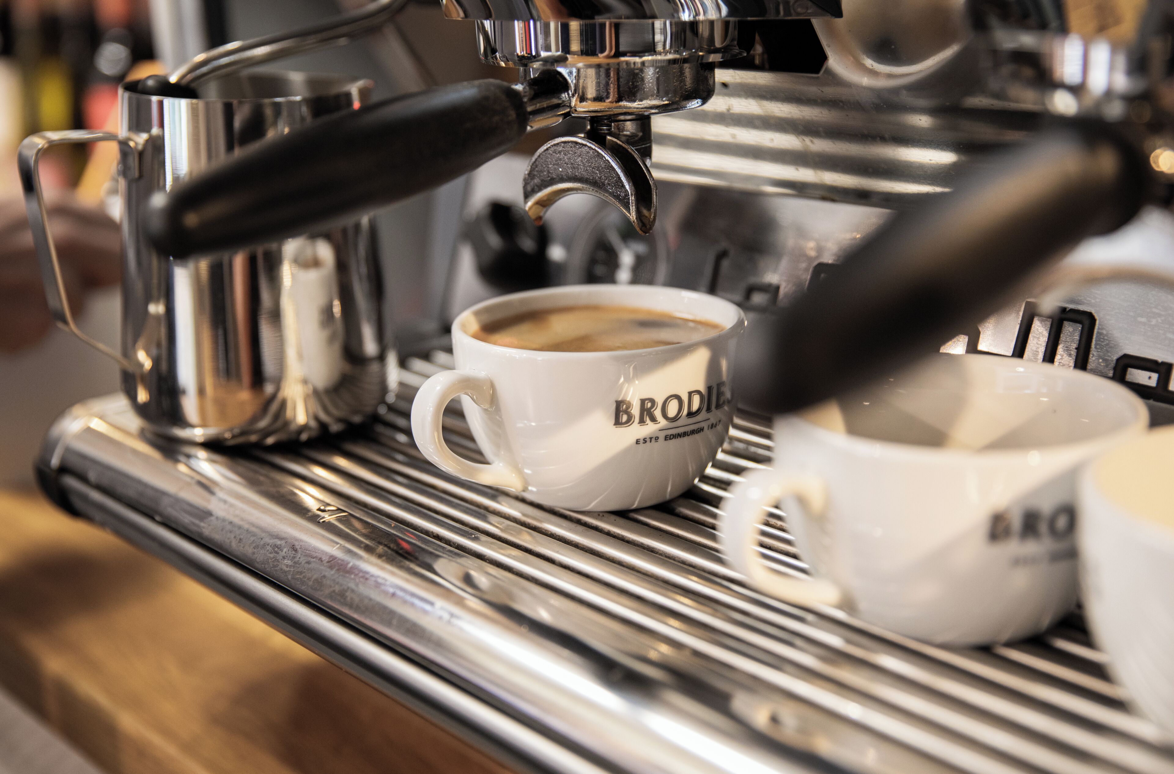 Una cafetera sirviendo café en una taza con la etiqueta 'BRODIE'.