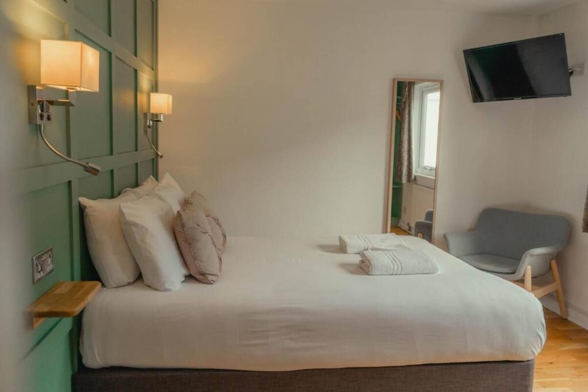 A neatly made bed with pillows and a folded towel, a bedside table, a chair, a mounted TV, and a mirror on the wall.