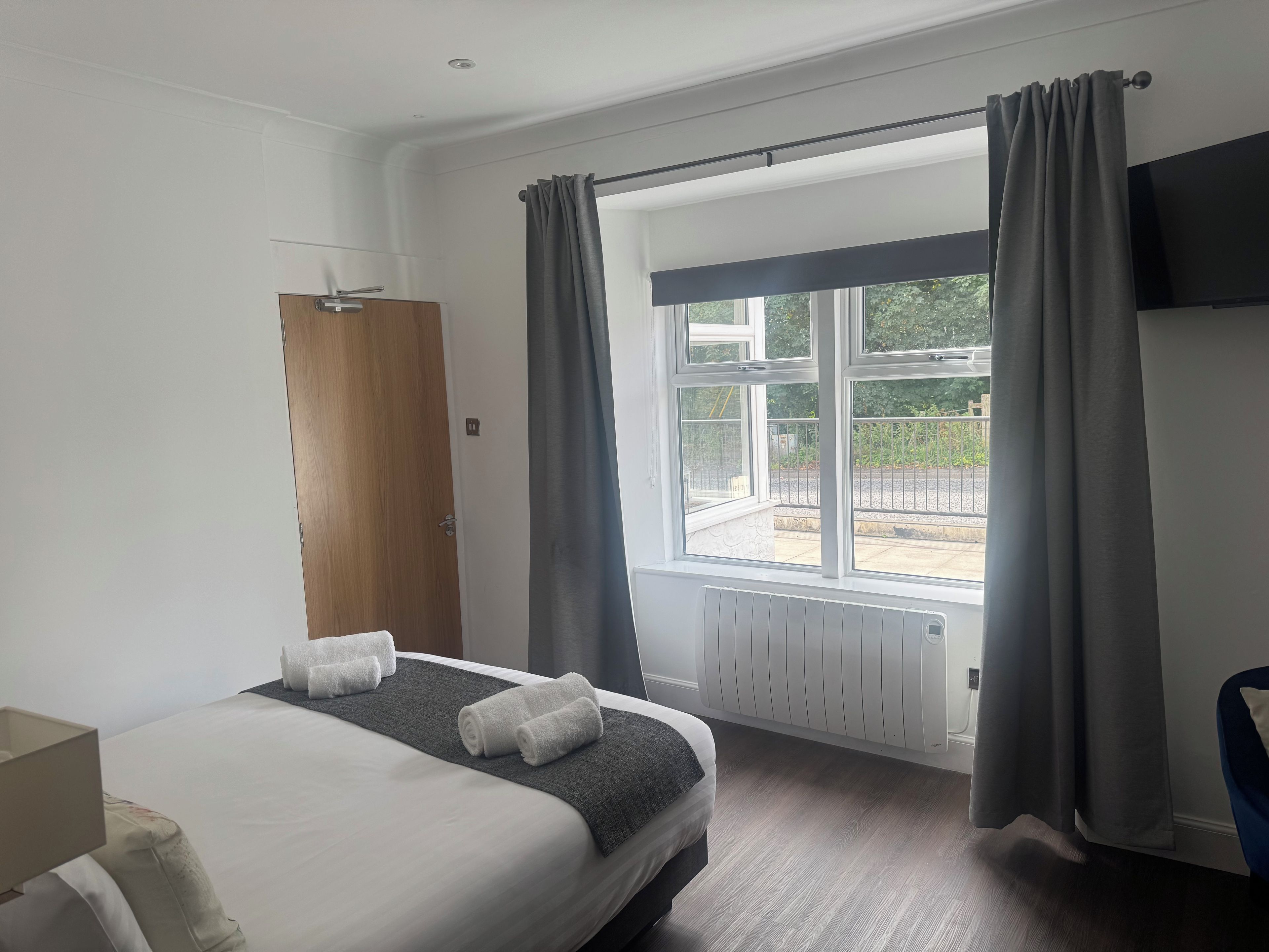 Ein Hotelzimmer mit Bett, Holztür, einem Fenster mit Vorhängen, einem Heizkörper und einem an der Wand befestigten Fernseher.