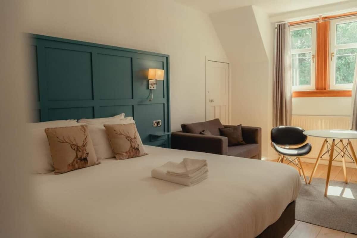 A hotel room with a large bed, a teal headboard, a desk with a chair, and a window with curtains.