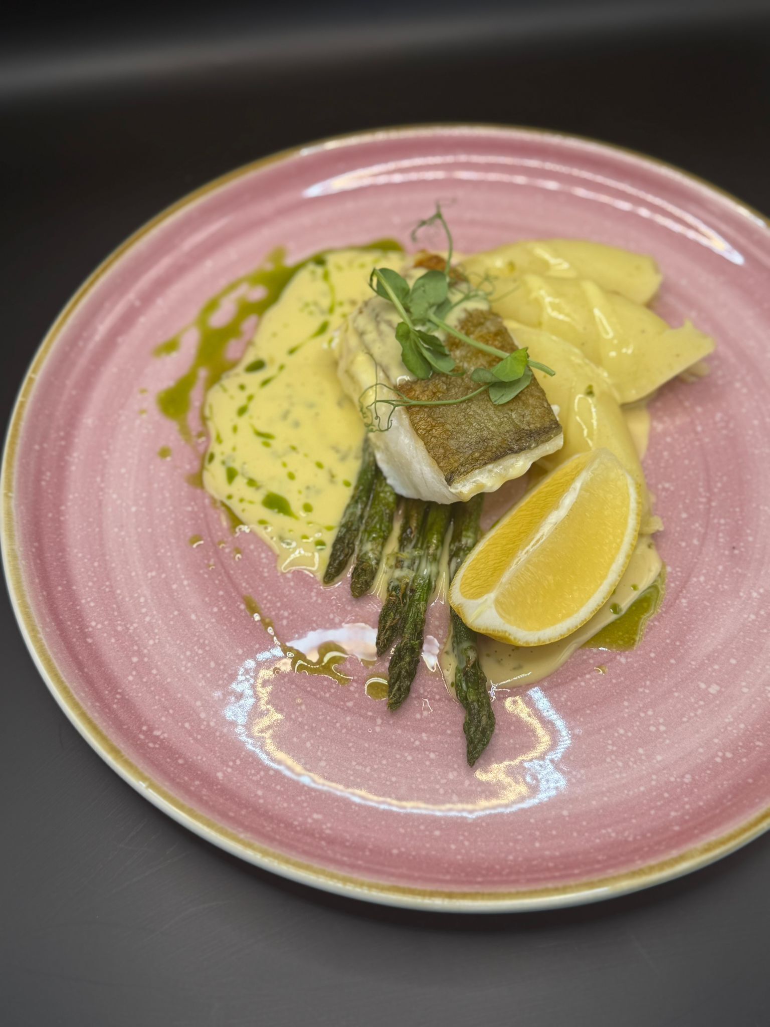 Un plato rosa con pescado, espárragos, limón y salsa.