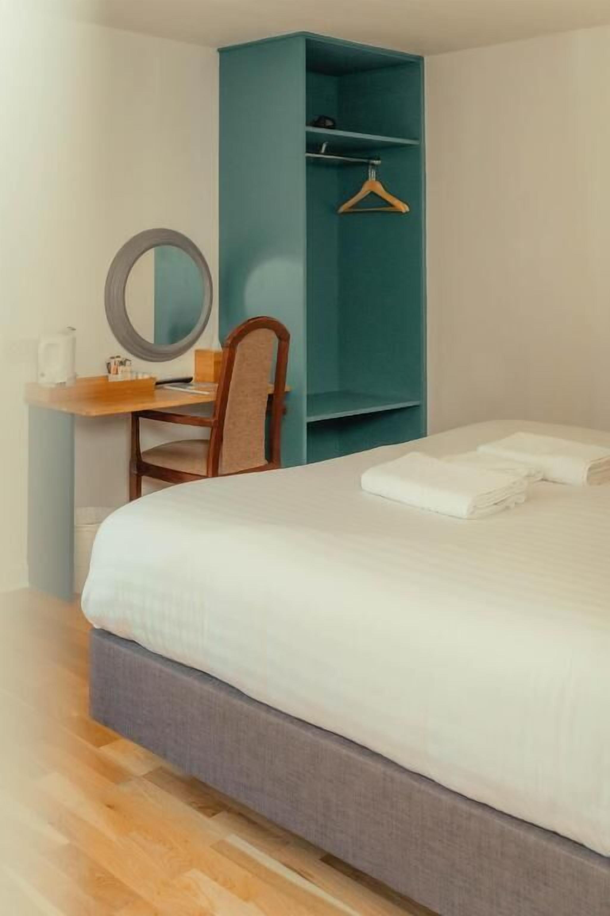 A hotel room with a bed, a desk with a chair, a mirror, and a teal storage unit.