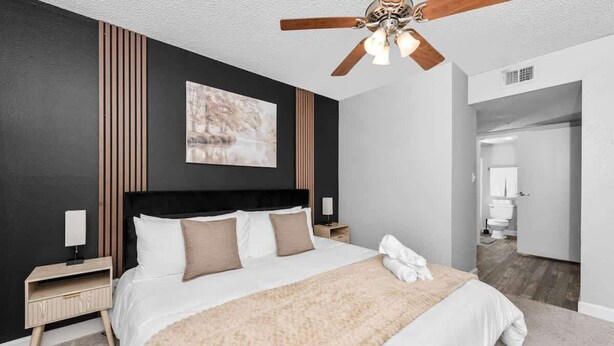 A modern bedroom with a large bed, a ceiling fan, a wooden panel wall, and a painting. - Snug 2BR NearASU Pool Grill, Pet Friendly, Parking (Tempe)