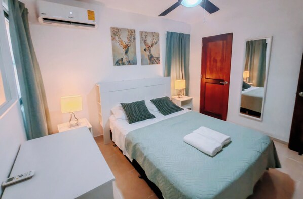 A hotel room with a bed, bedside lamps, a desk, an air conditioner, a ceiling fan, artwork, a mirror, and a door.
