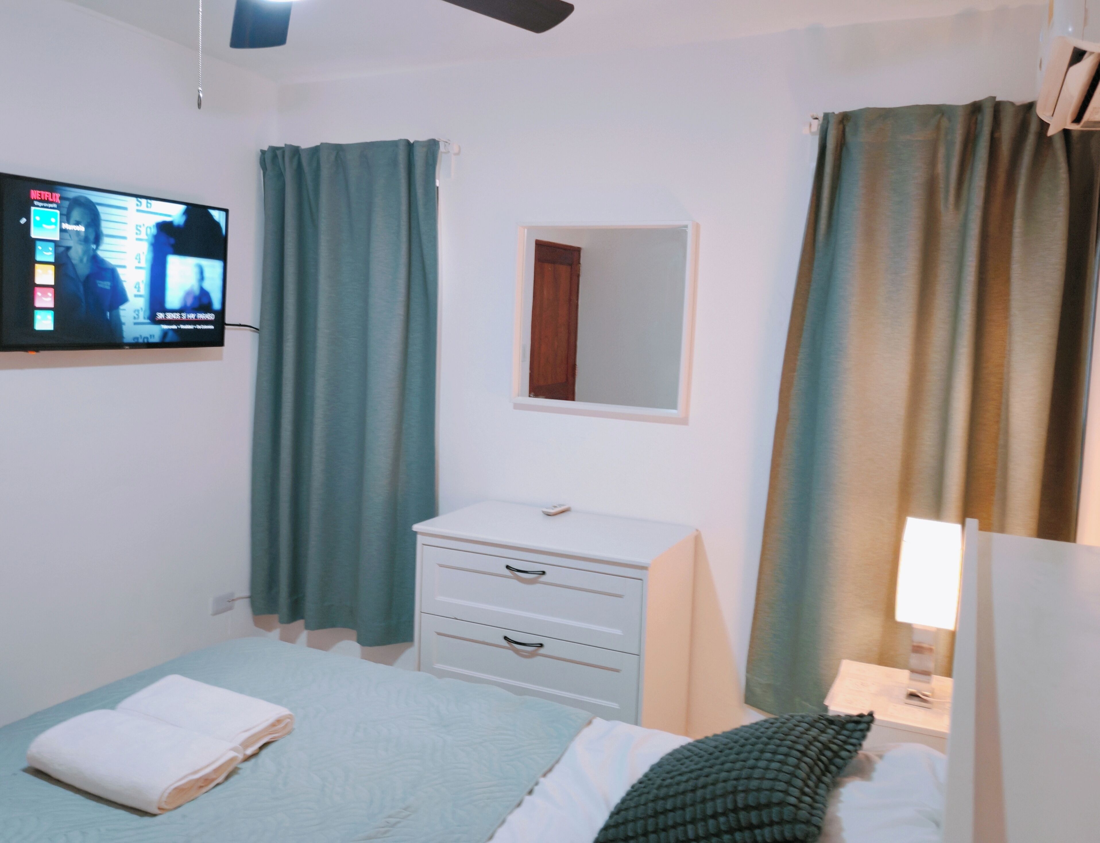 A bedroom with a bed, a bedside table with a lamp, a TV mounted on the wall, and a window with curtains.