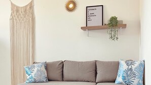 Uma sala de estar moderna com um sofá sectional cinza, uma mesa de centro de madeira, um tapete estampado, um painel de macramé na parede, uma decoração de parede em formato de sol e uma placa emoldurada numa prateleira.